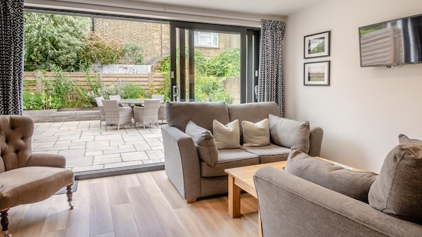 A spacious living area with a grey sofa, a wooden coffee table, and a patterned armchair. A large sliding glass door leads to an outdoor patio with a dining set and greenery. - 7 Bed in Aldeburgh (oc-midba) (Aldeburgh)