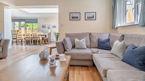 A spacious living room with a large beige sectional sofa, a wooden coffee table, and a dining area with wooden chairs and a table. - 7 Bed in Aldeburgh (oc-midba) (Aldeburgh)