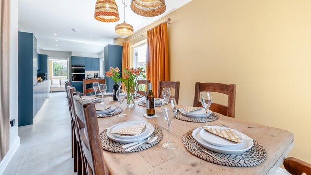 A dining area with a wooden table set for six, surrounded by chairs. There are woven placemats, white plates, and clear wine glasses. A vase with flowers is on the table, and the room has a modern design with blue walls and orange curtains. - 4 Bed in St. Peters (oc-h32665) (Broadstairs)