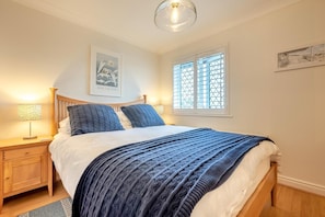 Ein Schlafzimmer mit einem Bett, einem Nachttisch, einer Lampe, einem Fenster mit Jalousien und einem Wandbild.