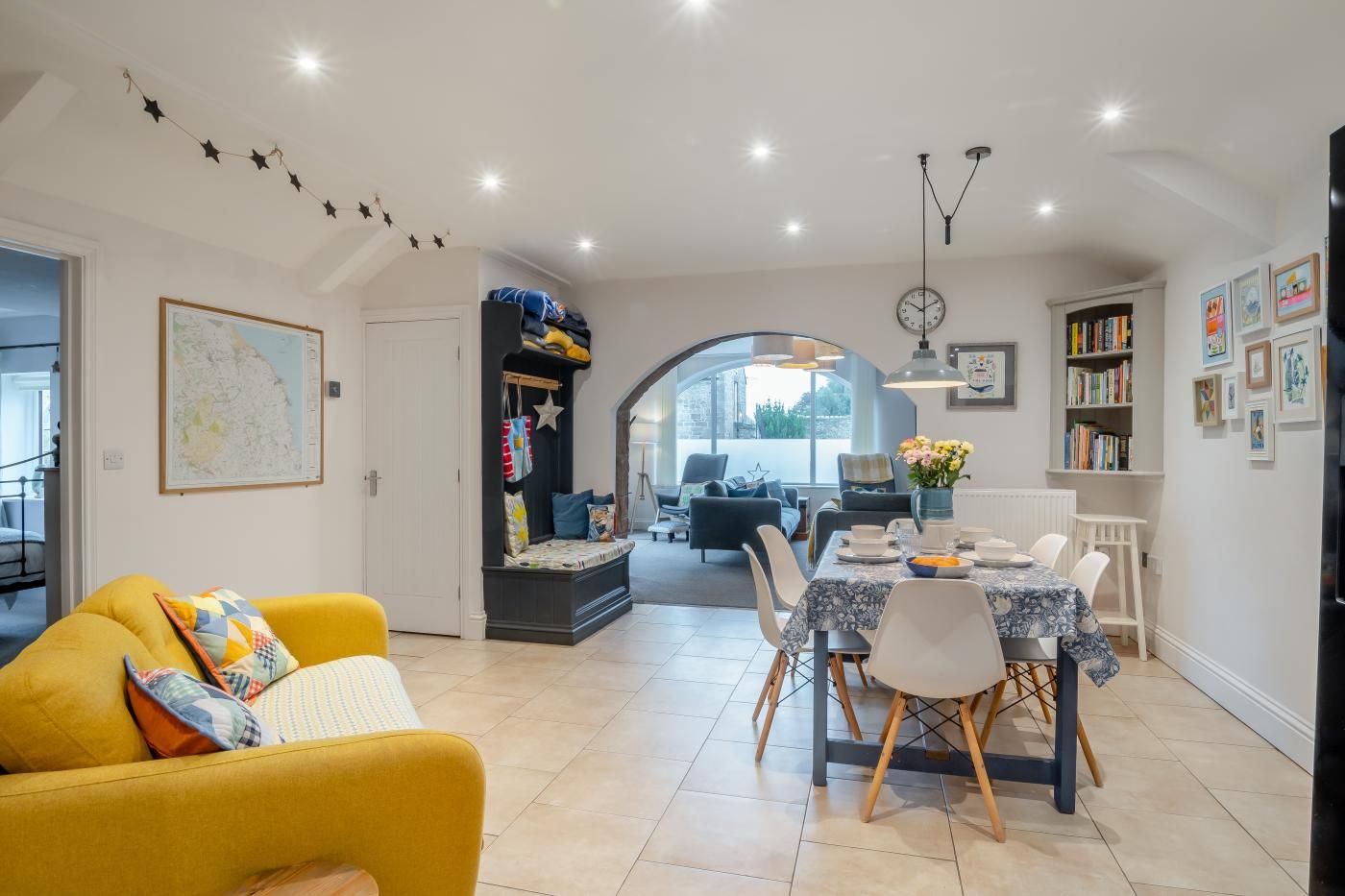 A modern living room with a yellow sofa, a dining area with a table set for four, and a kitchen visible in the background.