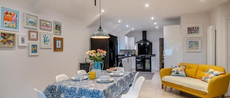 A dining area with a table set for two, a yellow sofa, and a kitchen in the background.