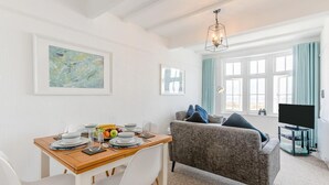 A dining area with a wooden table set for a meal, a grey sofa with blue cushions, and a television on a stand. - 2 Bed in St Ives (oc-pc055a) (St Ives)
