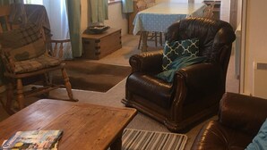 Ein gemütliches Wohnzimmer mit einem Holztisch, einer Ledersofa und einem Schaukelstuhl. Im Hintergrund sind ein Esstisch und Stühle zu sehen.