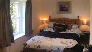 Ein Schlafzimmer mit einem Bett, zwei Nachttischlampen, einem Fenster mit Vorhängen, einem gerahmten Bild an der Wand und einer kleinen Pflanze auf dem Nachttisch.