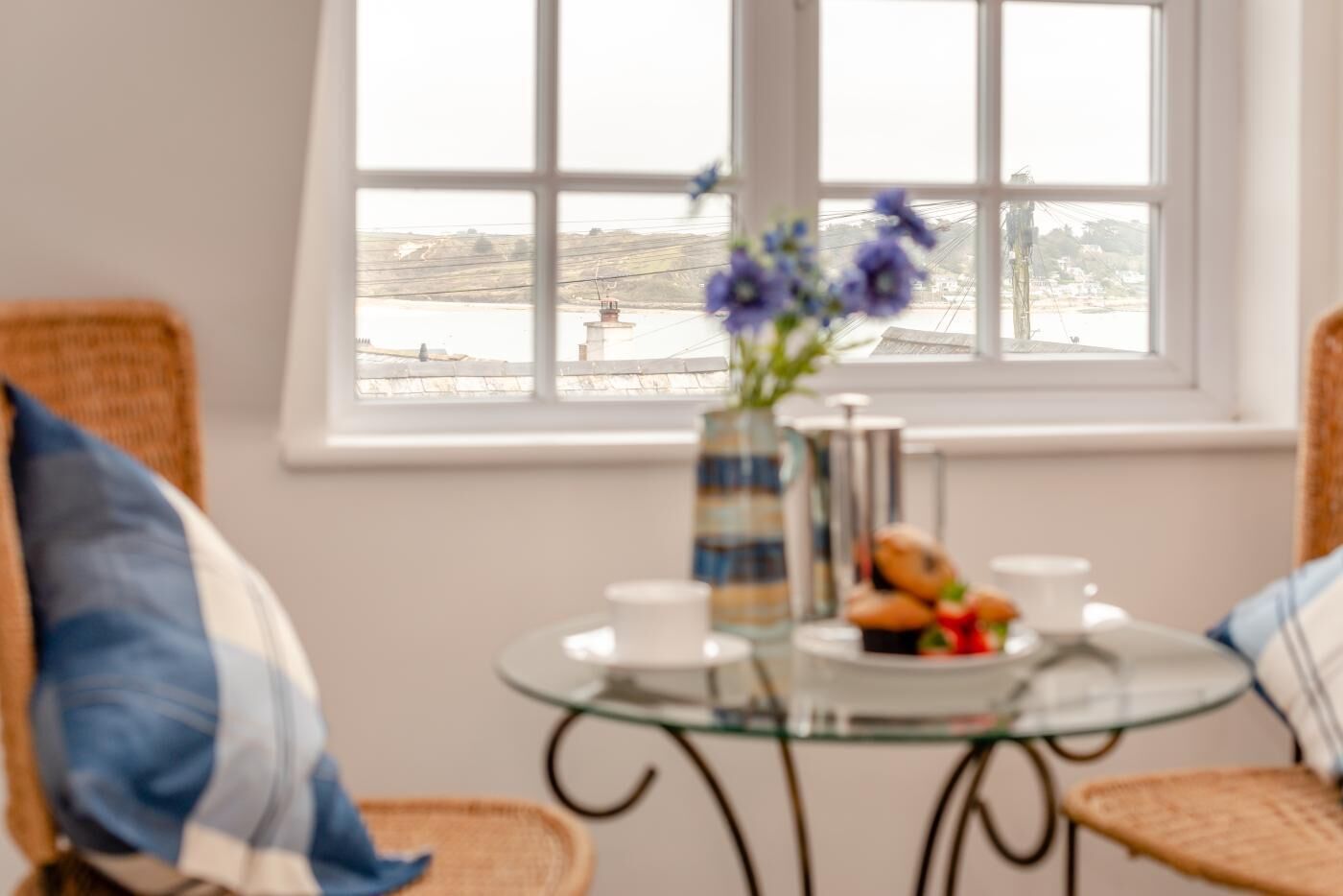 A room with a view of a body of water, a glass table with a vase of flowers, and a plate of pastries.