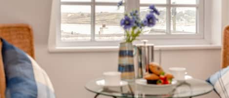 Une pièce avec vue sur un plan d’eau, une table en verre ornée d’un vase de fleurs et une assiette de pâtisseries.