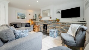 A modern living room with a grey sofa, a grey armchair, and a stone fireplace. A flat-screen TV is mounted on the wall above the fireplace. - 3 Bed in Padstow (oc-p00397) (Padstow)