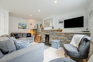 A modern living room with a grey sofa, a grey armchair, and a stone fireplace. A flat-screen TV is mounted on the wall above the fireplace. - 3 Bed in Padstow (oc-p00397) (Padstow)