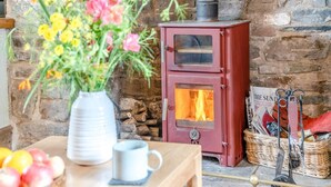 Une pièce rustique avec un mur en pierre, un poêle à bois rouge, un vase rempli de fleurs et une table sur laquelle on trouve des pommes et une tasse.