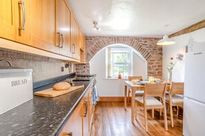 A kitchen with wooden cabinets, a countertop with a bread box, a stone wall, and a dining area with a table and chairs. - 2 Bed in Defynnog (oc-t32724) (Sennybridge)
