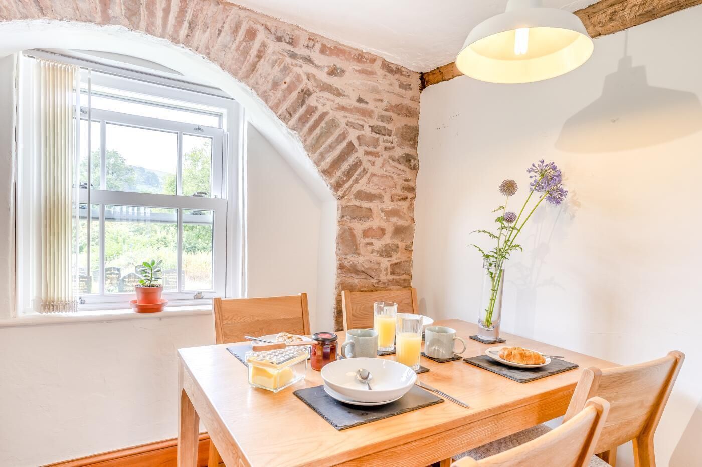 Une table de petit-déjeuner dressée avec une assiette, un bol, un verre de jus d’orange, un verre de lait, une tasse, un pot de confiture et une petite plante en pot.