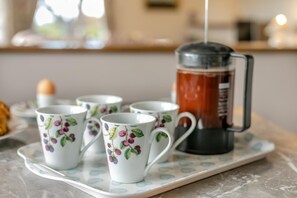 Un plateau avec quatre tasses blanches ornées d’un motif de fruits, une cafetière à piston noire et une assiette blanche à motifs bleus.