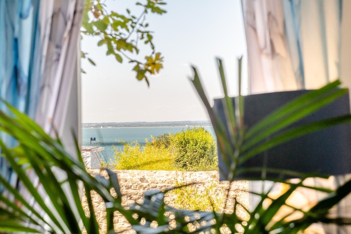 A view through an open window with a stone wall and greenery in the foreground, overlooking a body of water and distant land.
