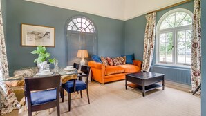 A room with a dining table set for four, an orange sofa, a dark coffee table, and a large window with curtains. - 1 Bed in Wymondham (oc-e32664) (Wymondham)