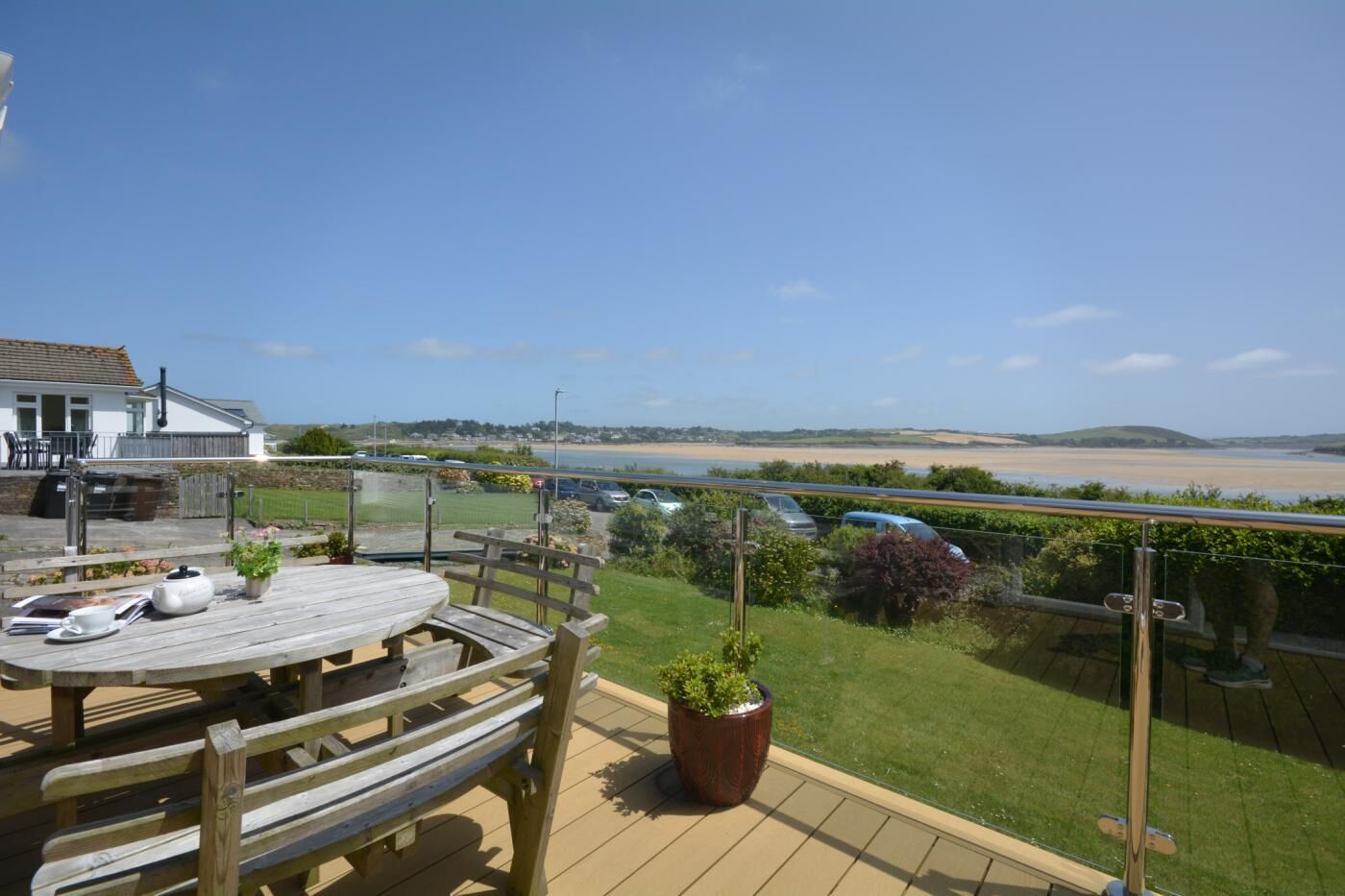 A wooden deck with a table and chairs, overlooking a beach and a parking area.