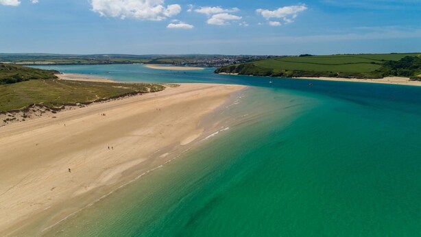 Eine Luftaufnahme einer Küstenlandschaft mit Sandstrand, klarem türkisblauem Wasser und grünen Hügeln im Hintergrund.