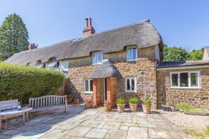 A stone house with a thatched roof, a wooden bench, and potted plants in the courtyard.