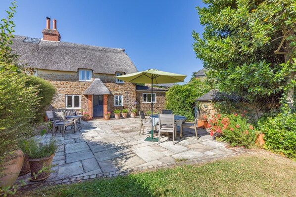 A stone house with a thatched roof, a patio with outdoor furniture, and a garden with potted plants and flowers.