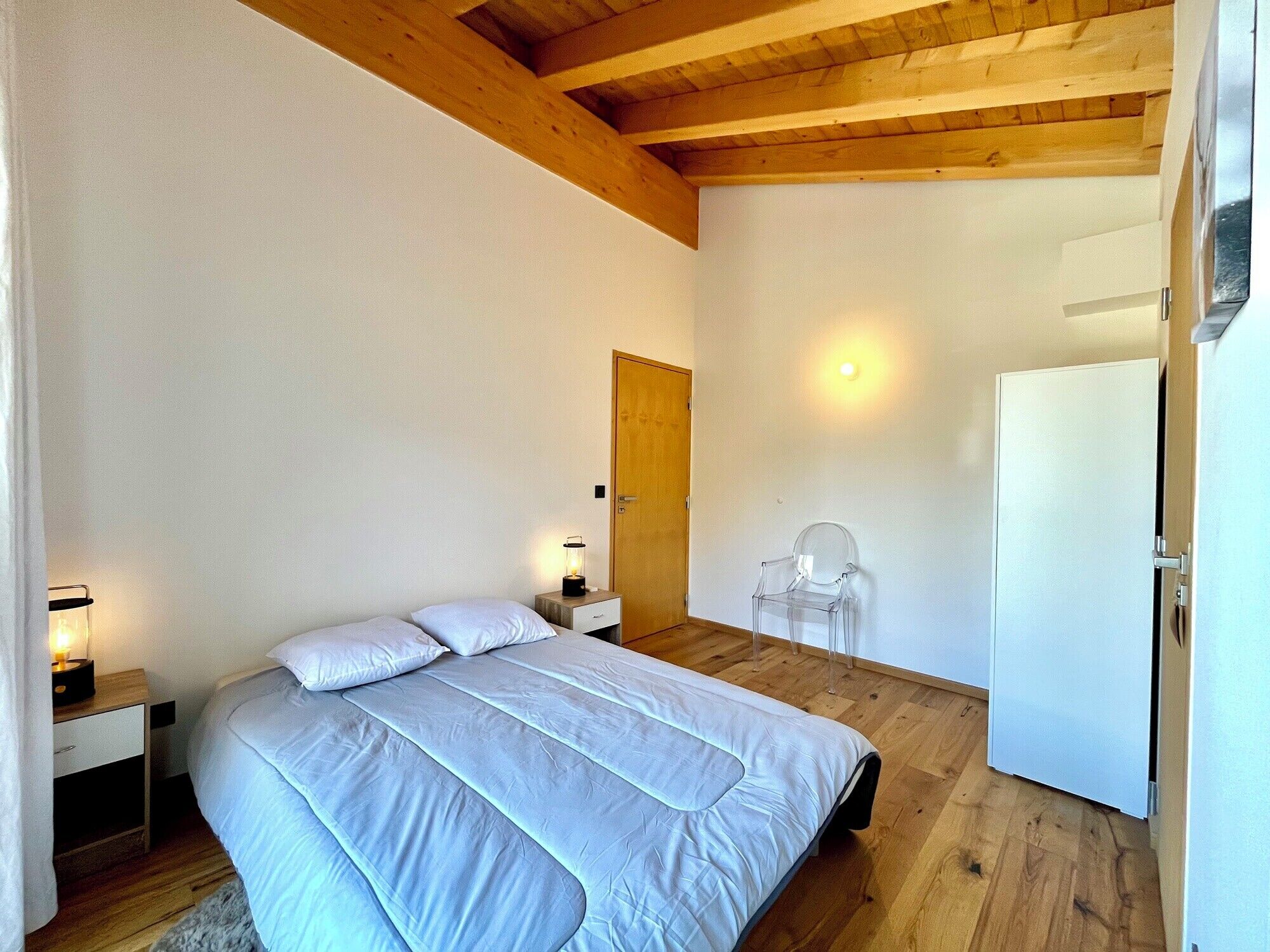 A bedroom with a wooden ceiling, a bed with white bedding, a nightstand with a lamp, a chair, and a white refrigerator.