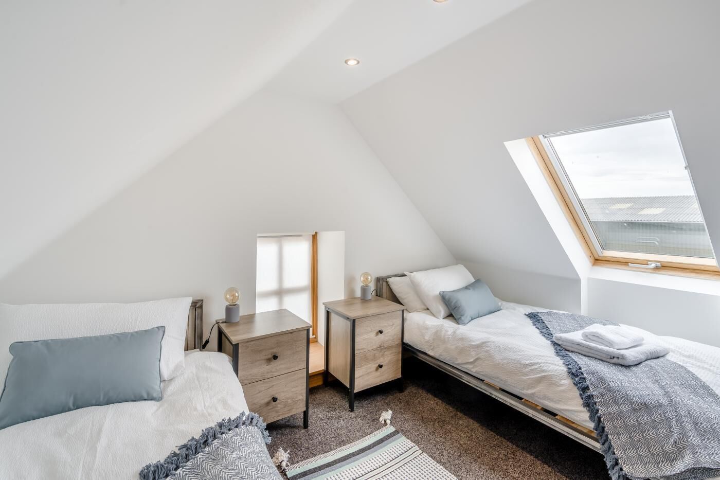 Un dormitorio moderno y luminoso con techo inclinado, tragaluz, dos camas, una mesita de noche y una pequeña mesa con lámpara.