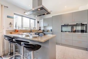 A modern kitchen with a central island, bar stools, stainless steel appliances, and a large window. - 3 Bed in Padstow (oc-p00329) (Padstow)
