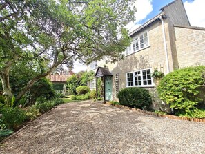 Une maison avec une allée gravillonnée, une porte verte et un jardin où poussent différentes plantes, ainsi qu’un arbre.