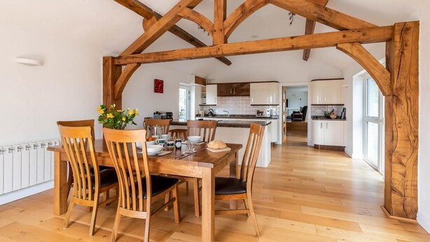 Un coin repas avec des chaises et une table en bois, une cuisine à l’arrière-plan et des poutres apparentes.