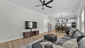 Una sala de estar moderna con televisor de pantalla plana, sofás oscuros, ventilador de techo y un comedor al fondo.