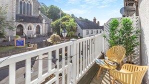 A balcony with a white railing, a yellow chair, and a table with a magazine and a cup. In the background, there is a church with a pointed steeple and a white building with a black roof. - 3 Bed in Beer (oc-mbcvw) (Beer)