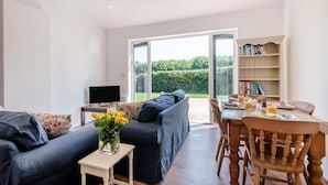 A modern living room with a sofa, a small table with a vase of flowers, a television, and a dining area with a table set for breakfast. - 2 Bed in Fressingfield (oc-j32677) (Fressingfield)