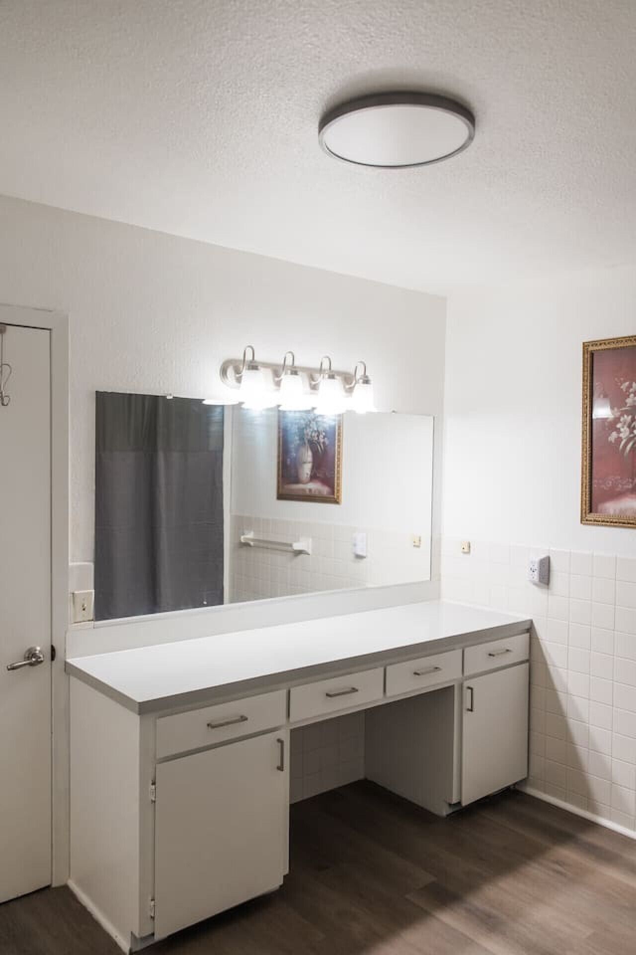 A bathroom with a white vanity, a mirror with lights, a framed picture, and white tiled walls.