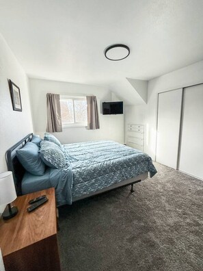 Un dormitorio con una cama, un televisor en la pared, una ventana con cortinas, un armario y suelo alfombrado.