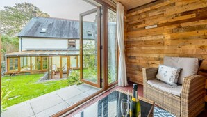 Une pièce chaleureuse avec une porte vitrée donnant sur un jardin, un canapé en osier et une table sur laquelle se trouvent une bouteille et des verres.