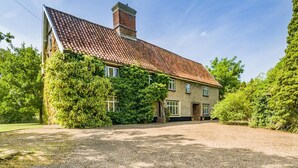 A two-story house with a gravel driveway, surrounded by lush greenery and trees.
