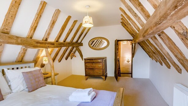 A room with a sloped ceiling, a bed with white linens, a wooden dresser, a round mirror, and a small table with a lamp.