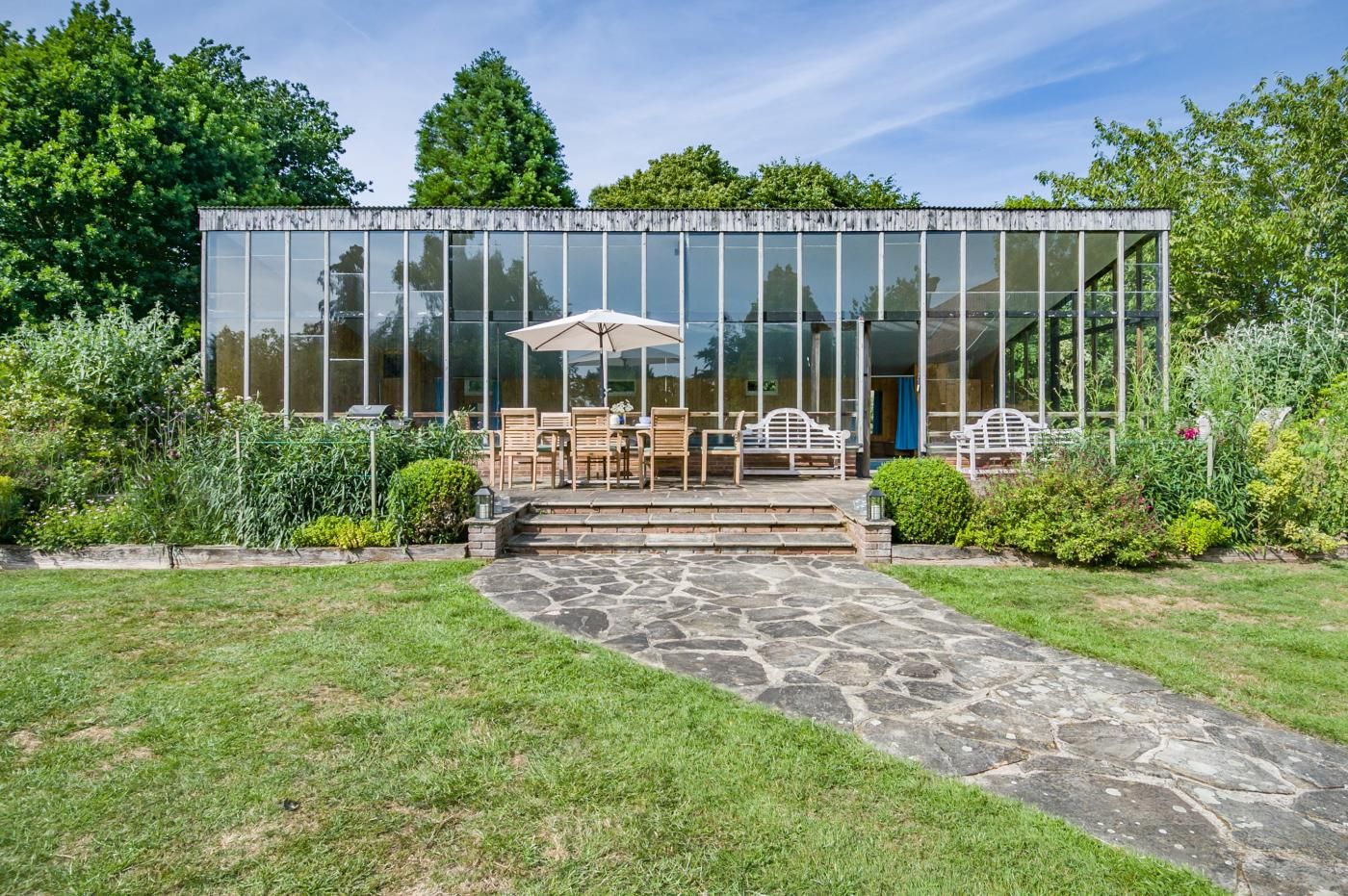 A modern glass house with a stone pathway leading to a patio with outdoor furniture.