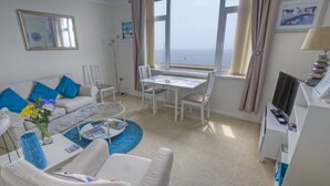 Une pièce comprenant un canapé, une table basse avec des magazines, une table à manger avec des chaises, un téléviseur et une vue sur la mer.