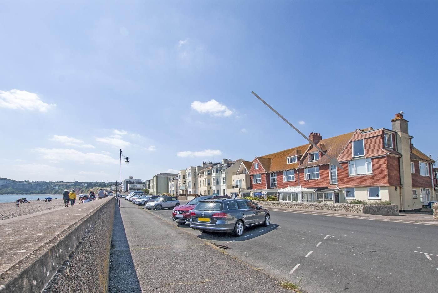 A coastal road with parked cars, a stone wall, and residential buildings.