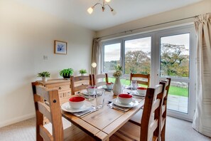 A dining area with a wooden table set for four, surrounded by chairs, with a view of a garden through large windows. - 4 Bed in Saundersfoot (oc-pw547a) (Saundersfoot)