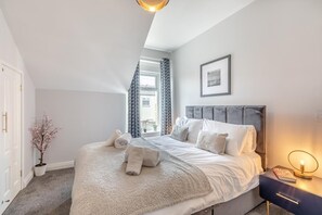 Ein Schlafzimmer mit Bett, Kissen, einem Nachttisch mit Lampe, einem Fenster mit Vorhängen und einem gerahmten Bild an der Wand.
