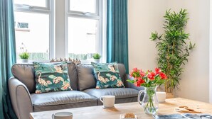 A modern living room with a grey sofa, floral cushions, a wooden coffee table, a vase with red flowers, and a tall green plant. - 3 Bed in Amble (oc-k32746) (Amble)
