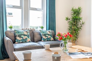 Ein modernes Wohnzimmer mit einem grauen Sofa, floralen Kissen, einem Holztisch, einer Vase mit roten Blumen und einer hohen, grünen Pflanze.