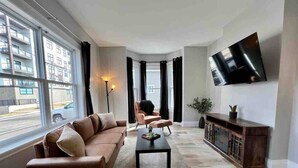 A living room with a sofa, a coffee table, a TV mounted on the wall, and a large window with curtains. - Downtown Oasis Moncton (Moncton)