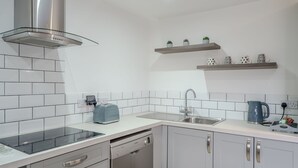 Eine moderne Küche mit einer Edelstahl-Dunstabzugshaube, weiß gefliestem Fliesenspiegel, einem Geschirrspüler und einer doppelten Spüle. Über der Spüle befinden sich zwei Regale mit Dekoration.