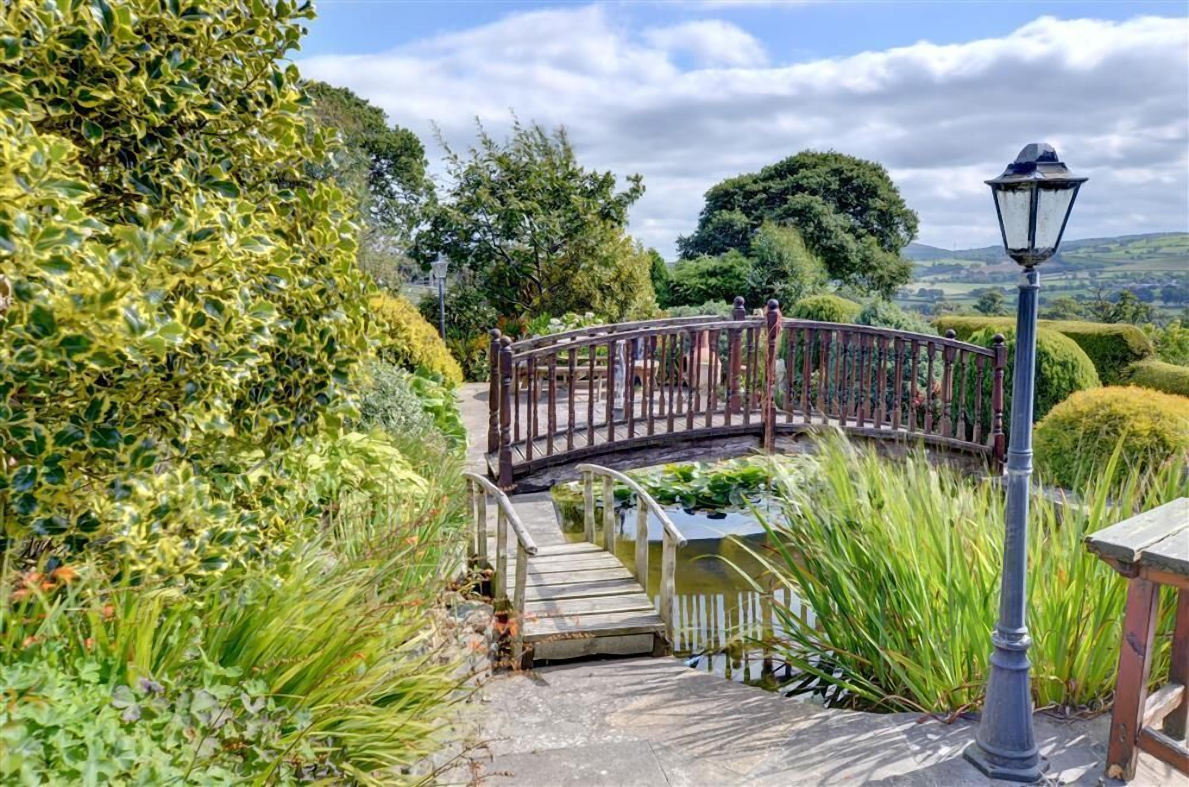 Eine Holzbrücke über einem Teich, umgeben von üppigem Grün und einer Laterne.