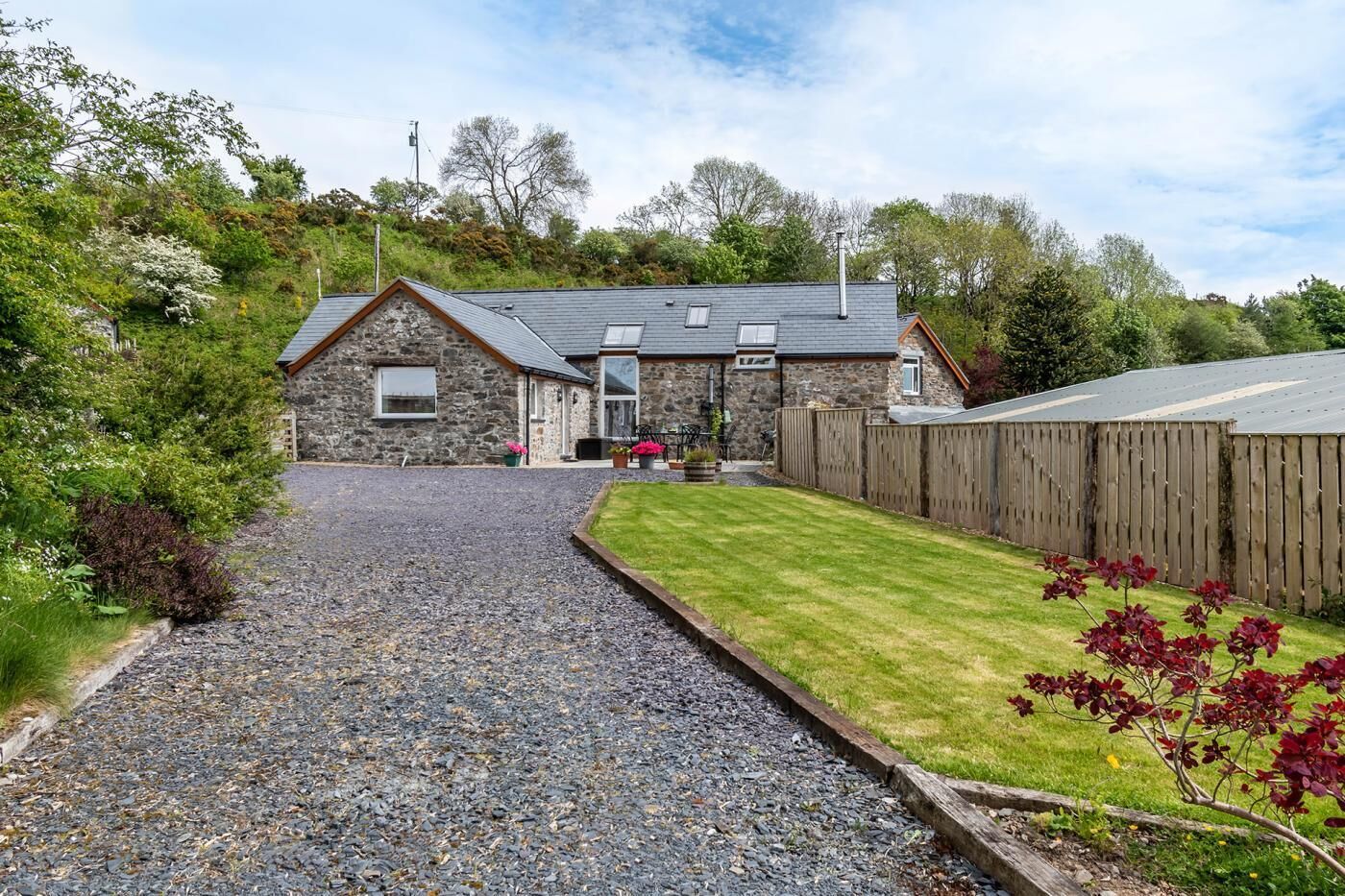 A stone house with a gravel driveway leading to a wooden fence and a grassy lawn.