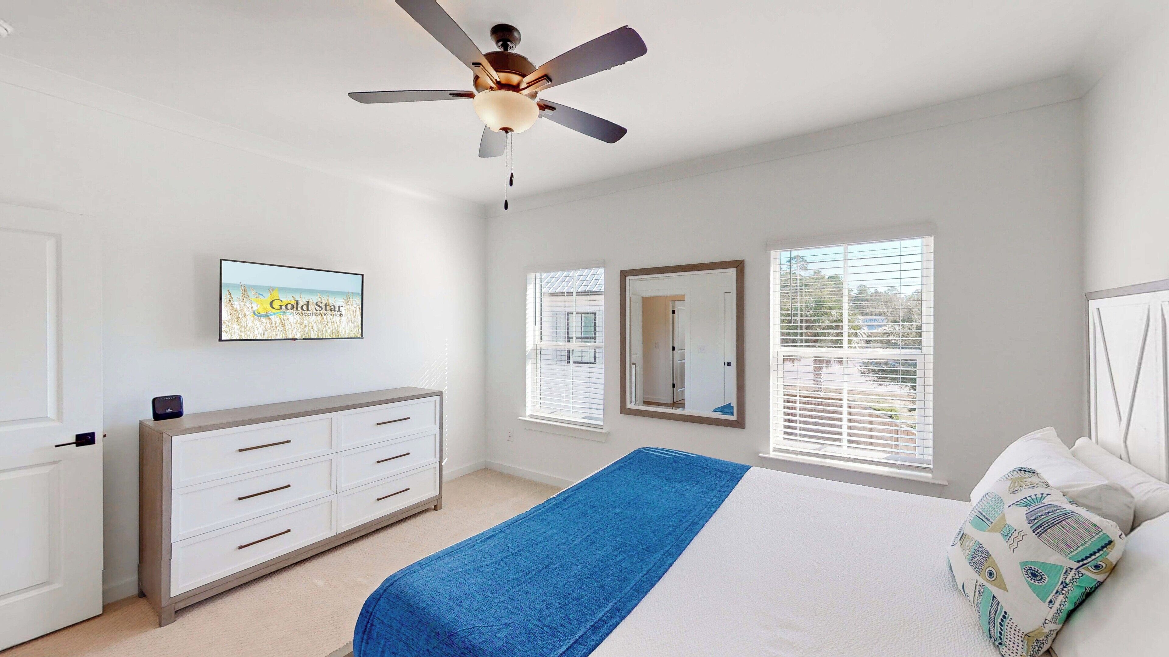 A bedroom with a bed, a ceiling fan, a dresser, a TV, and two windows with blinds.
