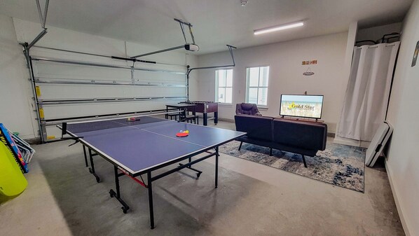 Une pièce équipée d’une table de ping-pong, d’un baby-foot, d’un canapé, d’un téléviseur diffusant une émission intitulée « Locations de logements pour personnes vaccinées » et d’une fenêtre avec des stores.
