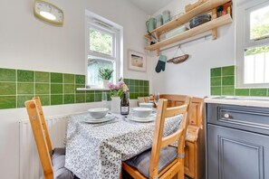 A kitchen with a dining table set for four, a window with a view, and a shelf with kitchenware. - 2 Bed in Saundersfoot (oc-pw948) (Saundersfoot)
